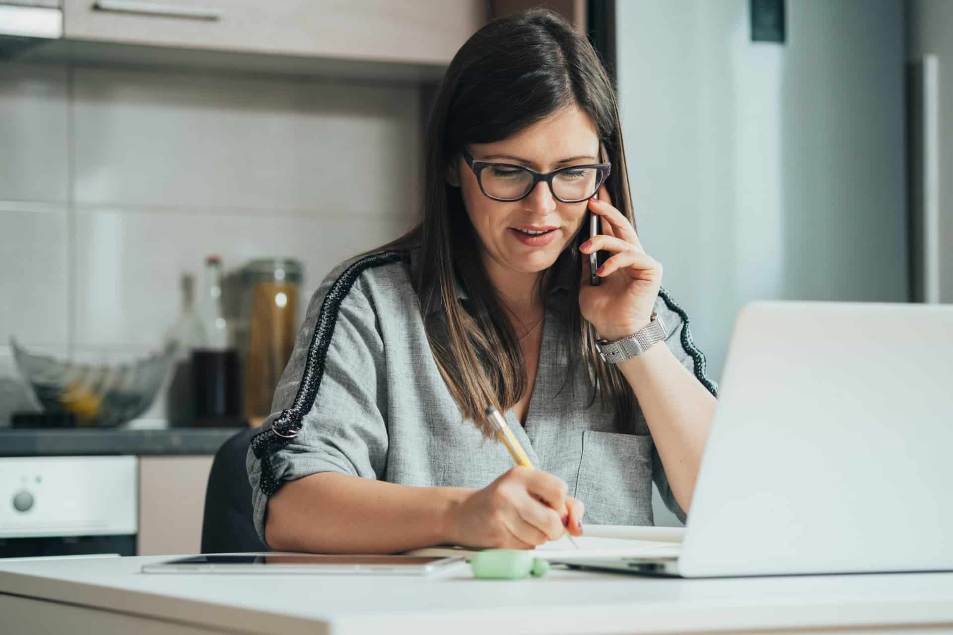 Smiling business woman talking on a mobile phone and writing notes in a notebook while sitting at the kitchen desk with digital tablet and laptop computer
